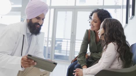 Médico-Sikh-Birracial-En-Turbante-Con-Tableta-Hablando-Con-Una-Chica-Con-Su-Madre-En-El-Hospital,-Cámara-Lenta