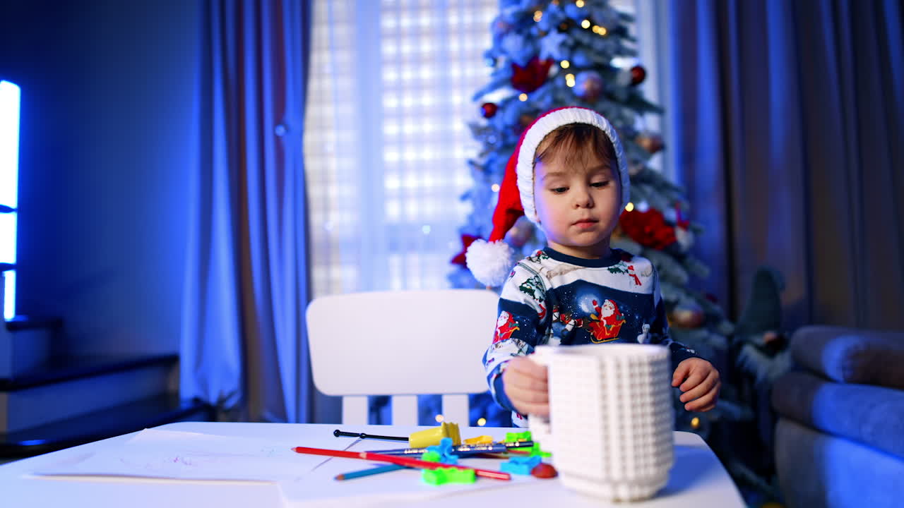 Peaceful baby boy in Santa cap stands holding a white big mug for pencils. Kid took away his pencils and drinks from a cup. Christmas tree at backdrop.