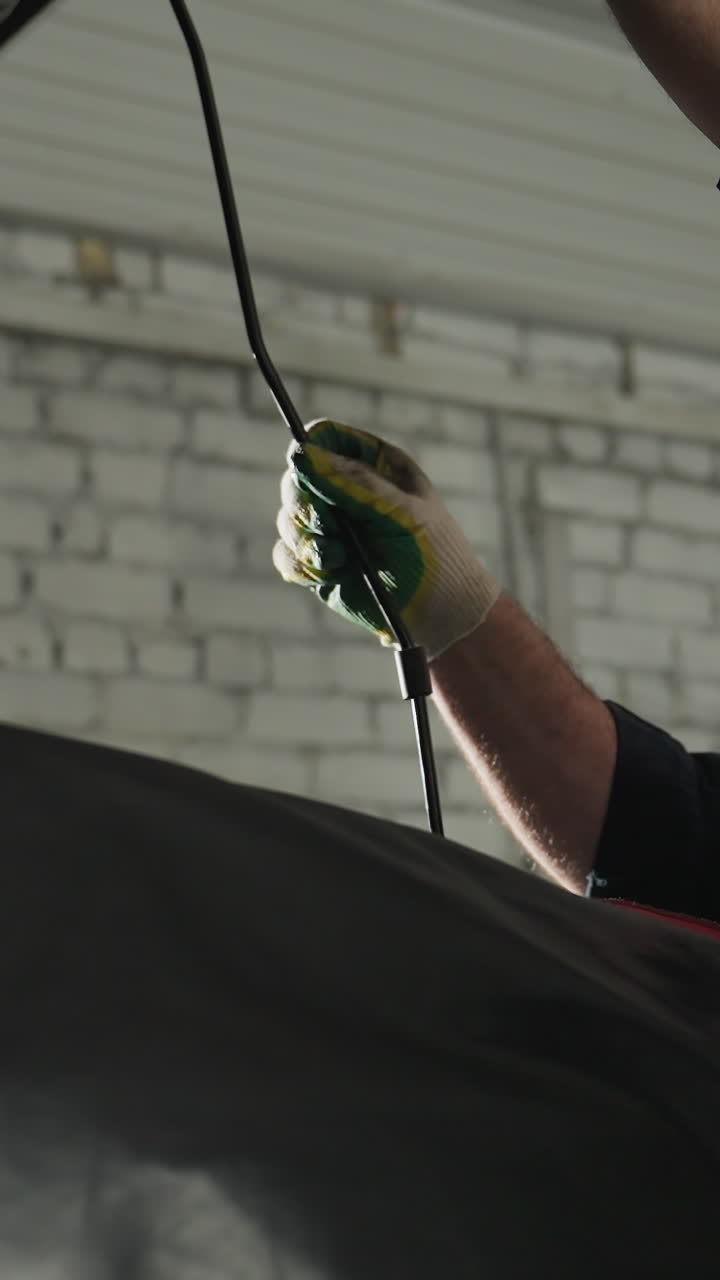 ingeniero de automóviles en uniforme azul habilmente utiliza una cuña para mantener abierto el capó de un coche negro en un taller mecánico bien iluminado, inspeccionando cuidadosamente los componentes del motor para el mantenimiento y la reparación