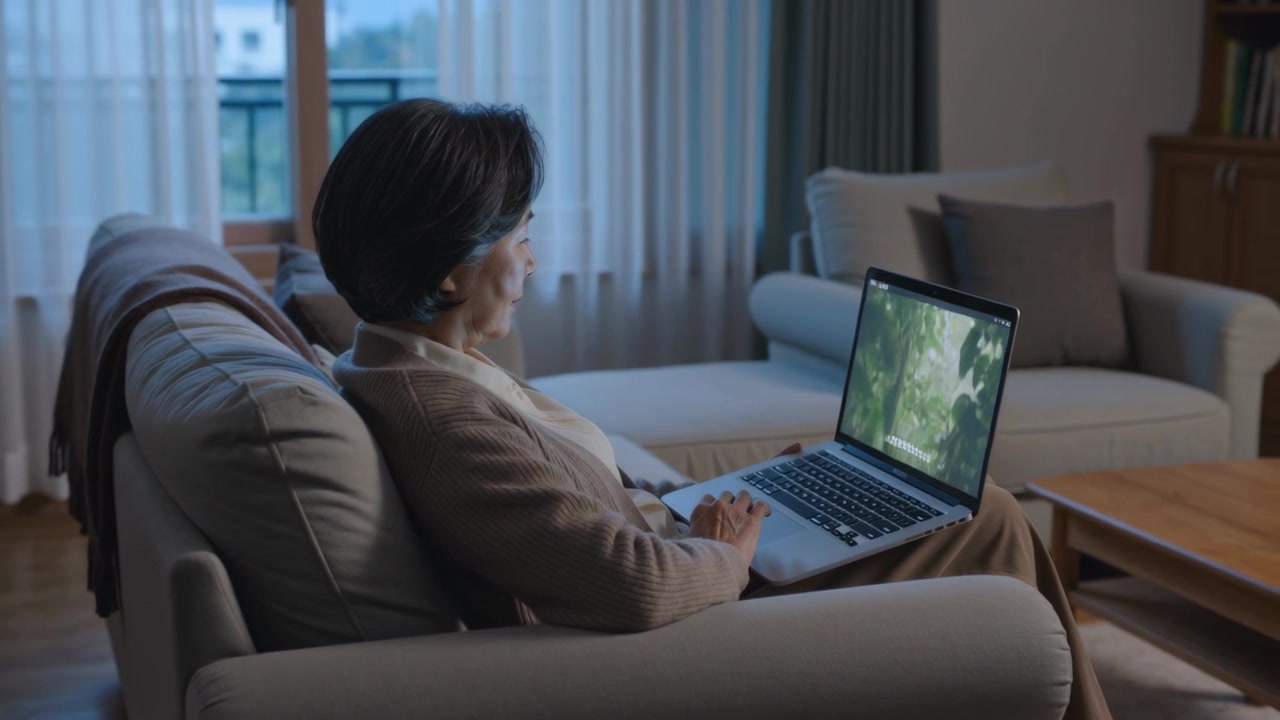 Watching Senior woman wearing cardigan adjusting on sofa at home while laptop playing nature scene