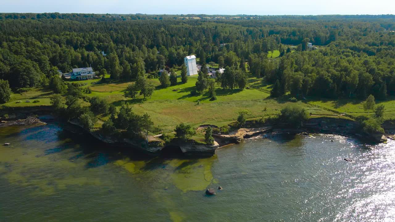 Gorgeous aerial drone footage orbiting and spinning around an old and tall white colored wooden lighthouse that is on sunny cliff bank ocean shoreline during summer time, with green grass, trees, sand