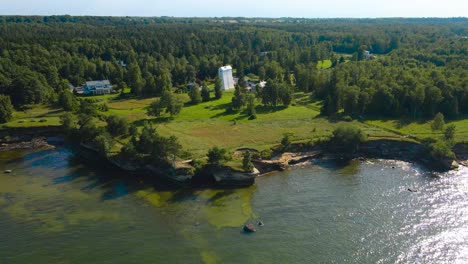 gorgeous aerial drone footage orbiting and spinning around an old and tall white colored wooden lighthouse that is on sunny cliff bank ocean shoreline during summer time, with green grass, trees, sand