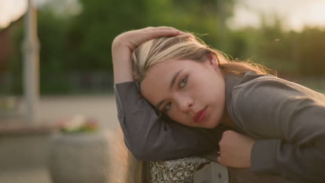 lady in grey clothing resting head on arm outdoors, gently touching her hair, lost in thought with a calm expression, eyes softly blinking, the background is slightly blur