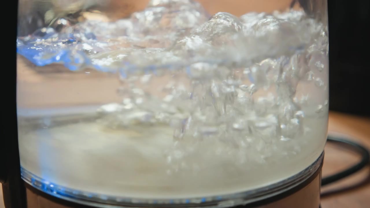 Vista ravvicinata estrema dal basso di un bollitore elettrico che riscalda l'acqua all'interno di una camera trasparente, con bolle dense, liquido in agitazione, vapore, illuminazione blu e superfici riflettenti in cucina