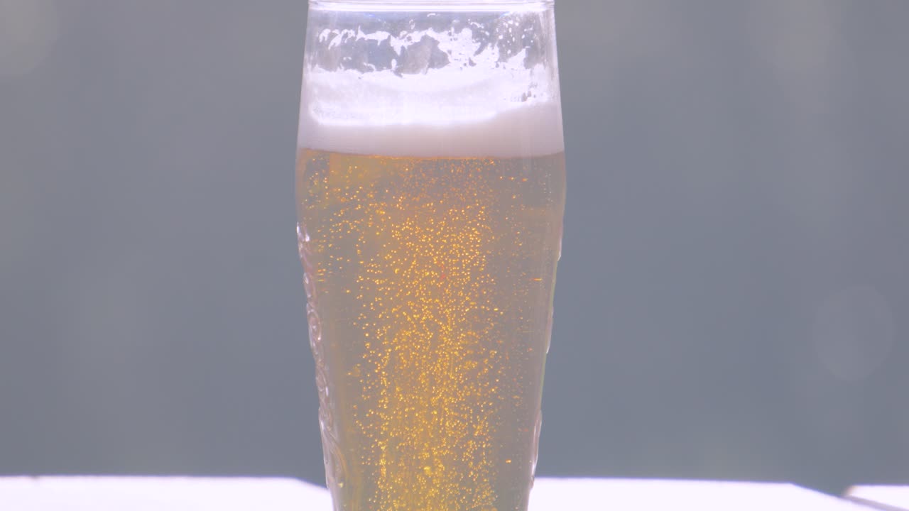 Bright Sunlight on Fresh Cool Lager Beer in Pint Glass with Bubbles Rising in Slow Motion on Wooden Picnic Table. Alcohol Beverage 4K.