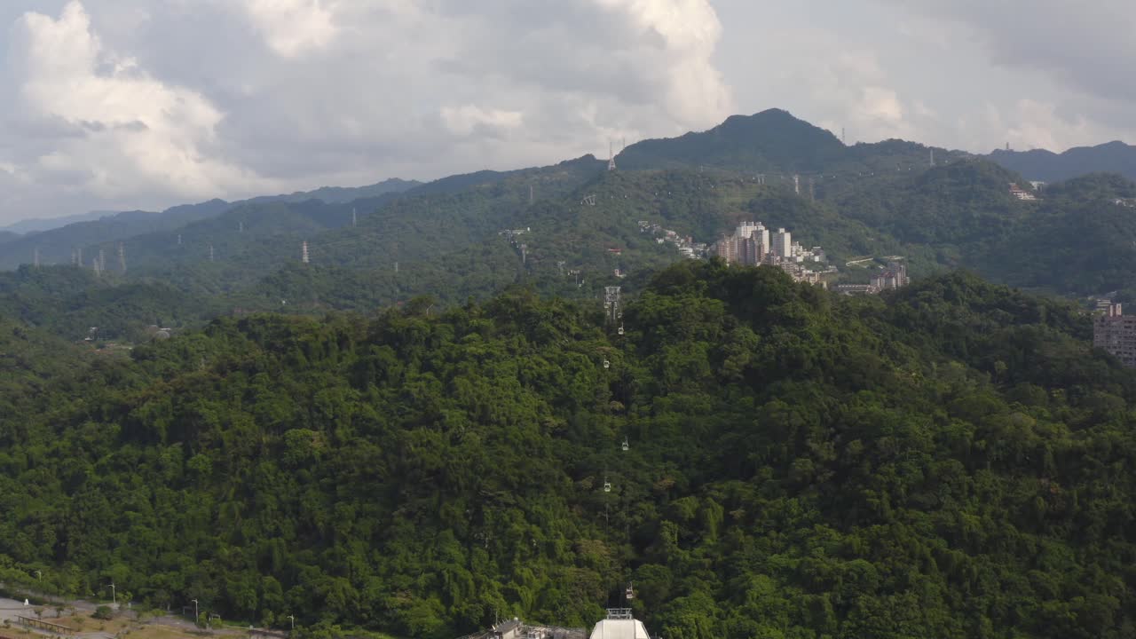 4K drone aerial zoom out shot of lush green mountain landscape with cable cars and distant city buildings under cloudy sky