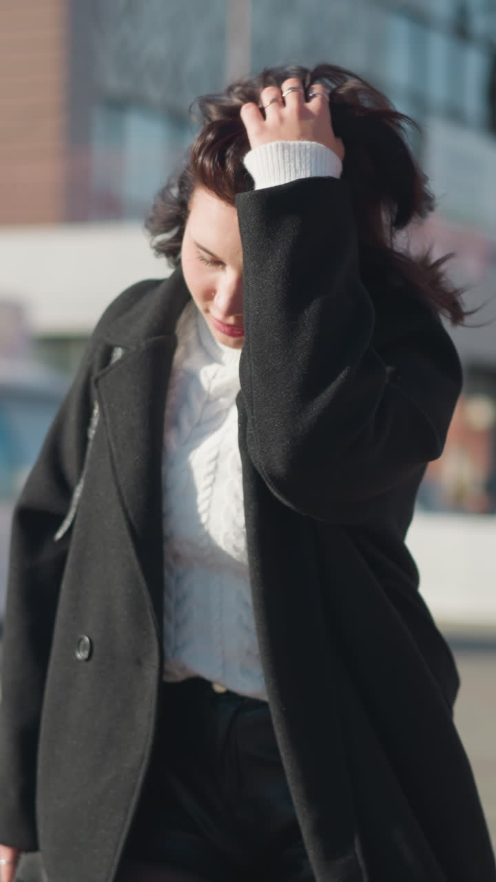 젊은 여성은 왼쪽으로 보면서 머리를 고, 색 케이블 뜨개질 스웨터와 검은색 코트를 입고, 현대적인 건물과 주차된 녹색 자전거를 배경으로 한 빛 도시 거리에서