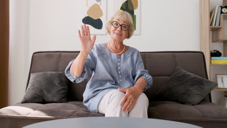 happy senior woman sitting on sofa in living room looking at camera and greeting during a video call at home