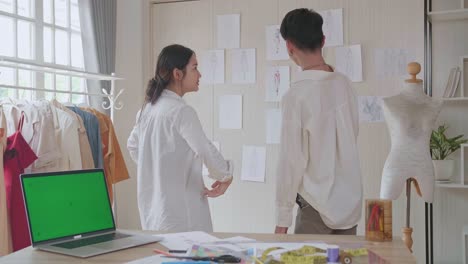 여성과 남성의 패션 디자이너는 녹색 화면 노트북 컴퓨터로 새로운 컬렉션의 옷 스케치를 선택하고 벽에 고정합니다.