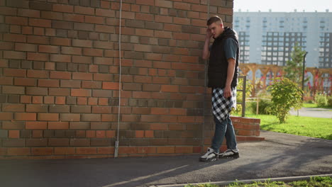 young boy in casual outfit walking beside building wall under sunlight reaches brick surface and rests before taking cigarette from ear and putting it into mouth with residential blocks blurred