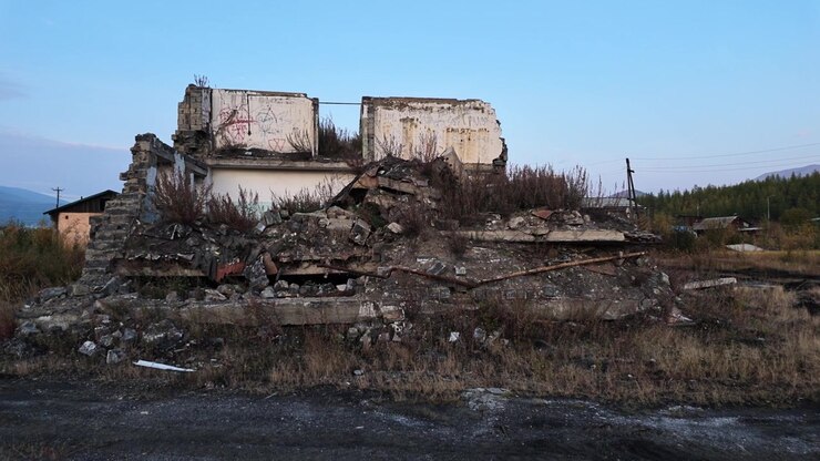 A static shot captures a dilapidated building with collapsed walls, covered in graffiti and surrounded by rubble and dry grass.