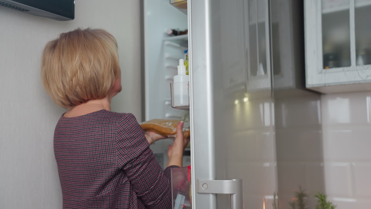 여성은 냉장고 안에 신선한 구운 반죽을 넣고, 다른 물건과 함께 음식을 조심스럽게 보관합니다. 현대적인 부 설정, 스테인리스 스 냉장기, 내부의 반사