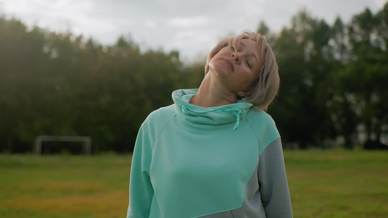Instructor wearing mint tracksuit performing head tilts exercise in open grassy field under soft cloudy sky during peaceful morning workout session focusing on flexibility posture correction