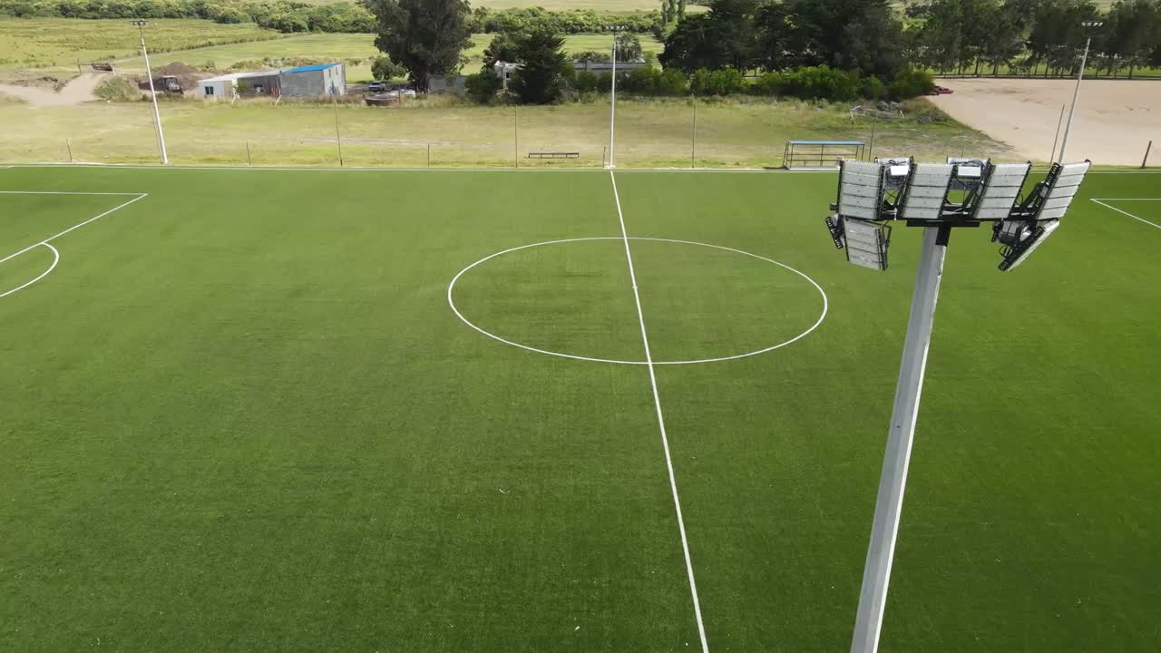 Rotating aerial perspective revealing empty soccer field with artificial green turf, showcasing stadium infrastructure and geometric landscape design