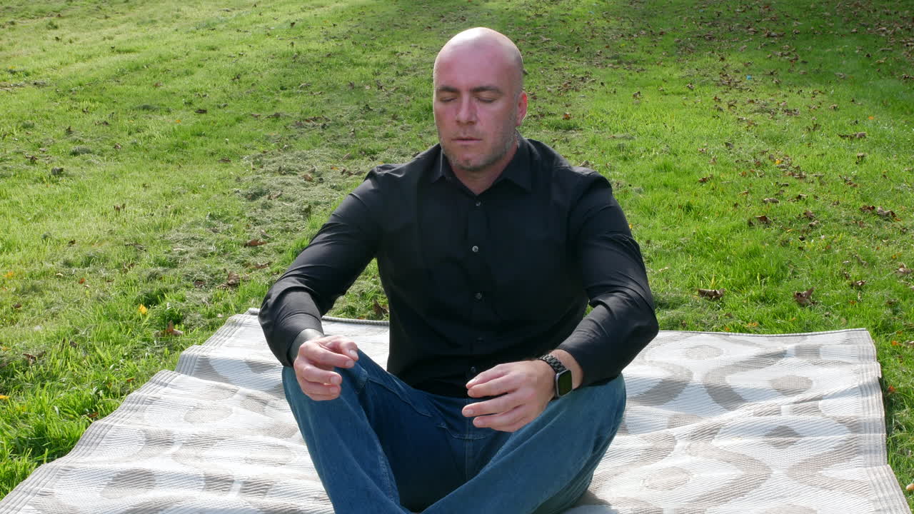 A man sitting in a grass field in a garden meditating in the yoga position on a mat