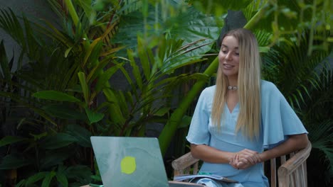 a girl in a blue dress remotely online working behind a laptop and looking into the screen the backyard with green plants