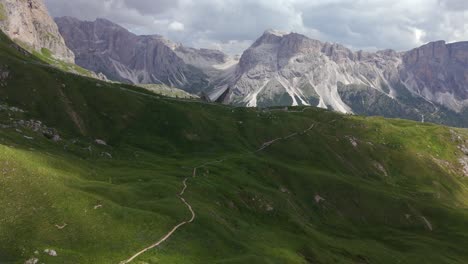 aerial view of a winding hiking trail cutting through the lush green valleys of the dolomites, surrounded by towering, rugged mountains under a dramatic sky