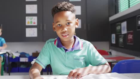 Portrait-of-happy-biracial-schoolboy-at-desk-in-school-classroom