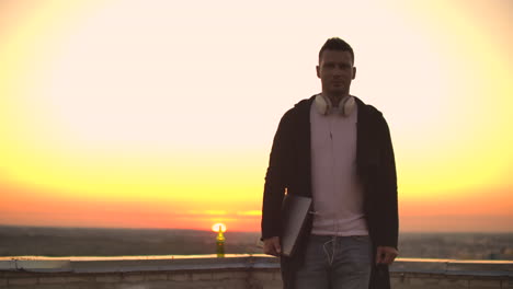 a man walks on the roof in a hoodie with a laptop in his hands to meet the camera.