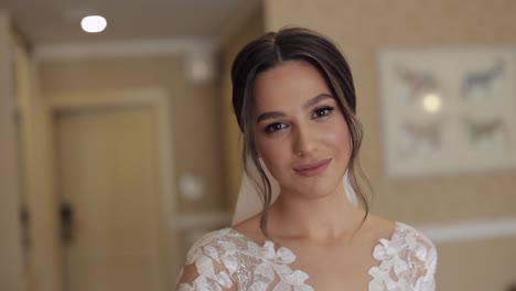 Lovely-bride-in-white-wedding-dress-and-veil-stay-at-home-in-living-room,-looking-at-camera,-smiling