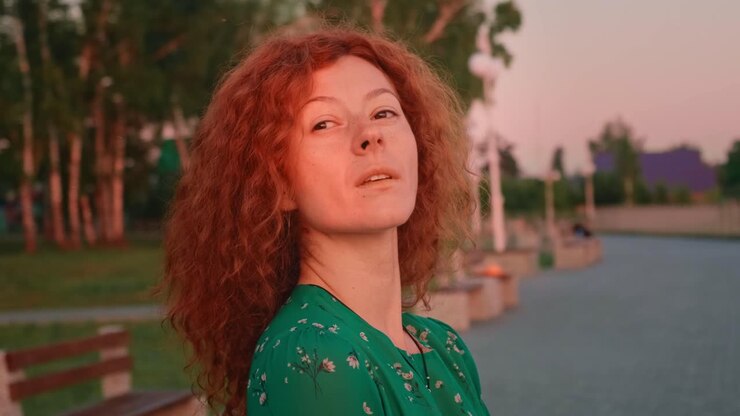 A Serene Evening Reflection: A Woman with Curly Red Hair Gazes Thoughtfully Towards the Horizon Amidst a Tranquil Park Setting at Dusk