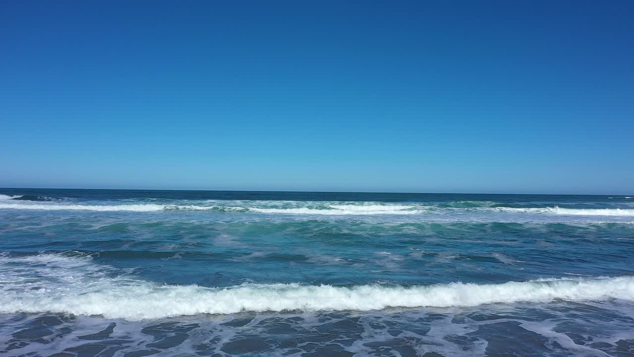 Aerial shot over waves slowly crashing with nothing but blue skies on the horizon