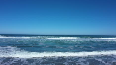 aerial shot over waves slowly crashing with nothing but blue skies on the horizon