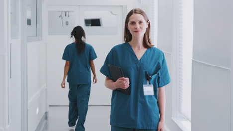 portrait of smiling female doctor wearing scrubs in busy hospital corridor holding digital tablet