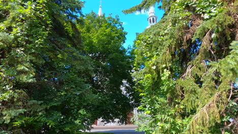 Der-Dolly-Bewegt-Sich-Zwischen-Bäumen-Hindurch-In-Richtung-Der-Wunderschönen-Kirche,-Kippt-Nach-Oben-Und-Gibt-Den-Blick-Auf-Die-Außenfassade-Des-Hohen-Gebäudes-Und-Den-Blauen-Himmel-Frei