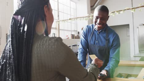 african american woman paying happy male barista by credit card at coffee shop, slow motion