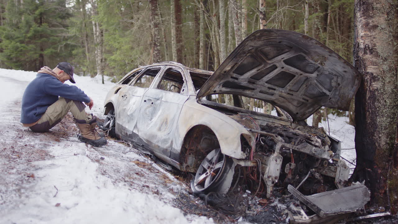 Despondent man sits next to wrecked and burnt out vehicle on snowy roadside