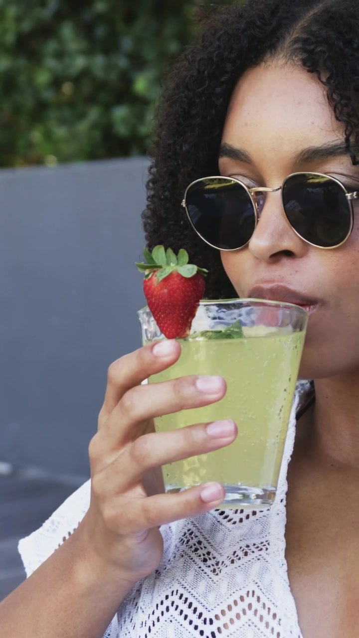 Video verticale: Donna in top bianco ispeziona il bicchiere, lo solleva per assaggiare, sorseggiando a bordo piscina, con fragola.