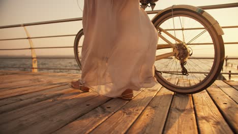 Close-up-shot-of-a-girl-in-a-long-white-light-dress-who-wriggles-in-the-wind-and-walks-near-her-bicycle-along-the-beach,-which-is-covered-with-boards-along-the-sea-at-Sunrise-in-summer