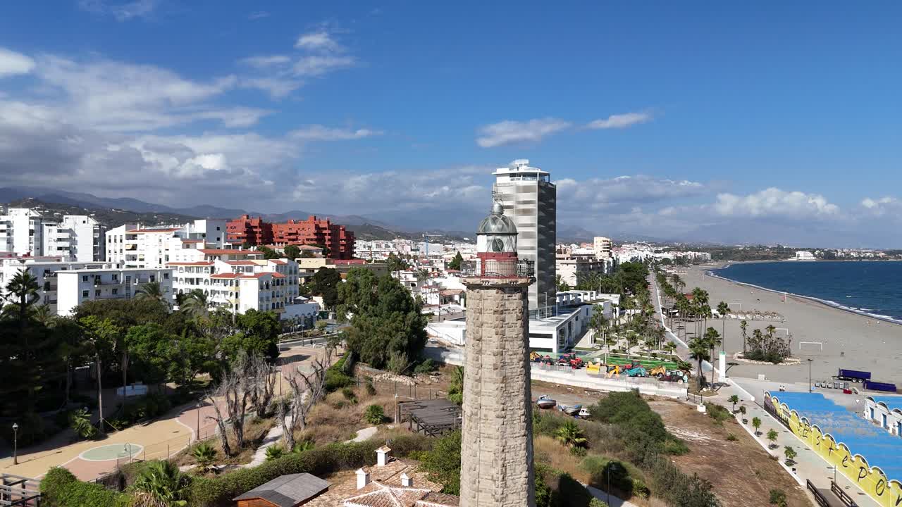 Drone tilt up shot of historic lighthouse near beach in Estepona with surrounding modern buildings trees coastline and blue sky showing contrast between heritage and development