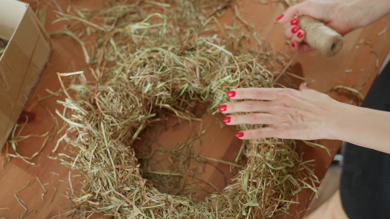 Hands with red nails shaping hay wreath on wooden table using twine for crafting natural rustic decoration, creating eco-friendly handmade accessory with straw material in creative workspace