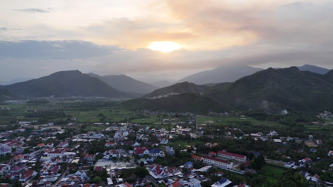 Sun sets above peaceful valley town in Rhodope Mountains with rolling green hills, distant farmlands and soft clouds casting golden glow over scenic Eastern European countryside