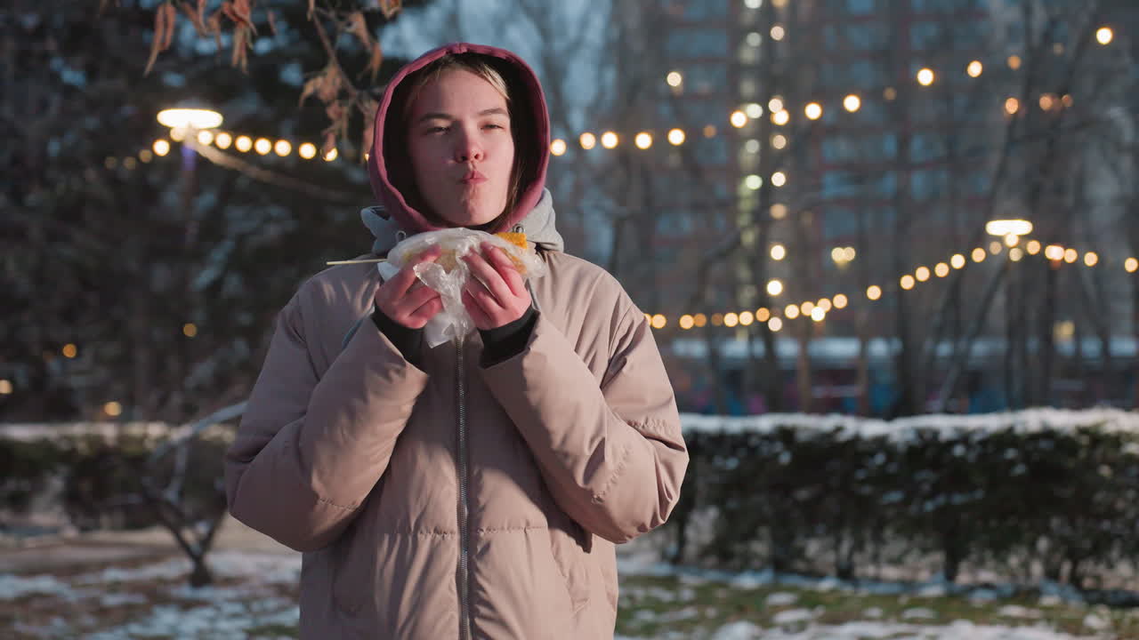야외에 서있는 젊은 여성, 막대기에 옥수수를 즐기고, 공원 설정에서 겨울 코트를 입고 배경에 보케 조명, 땅에 눈