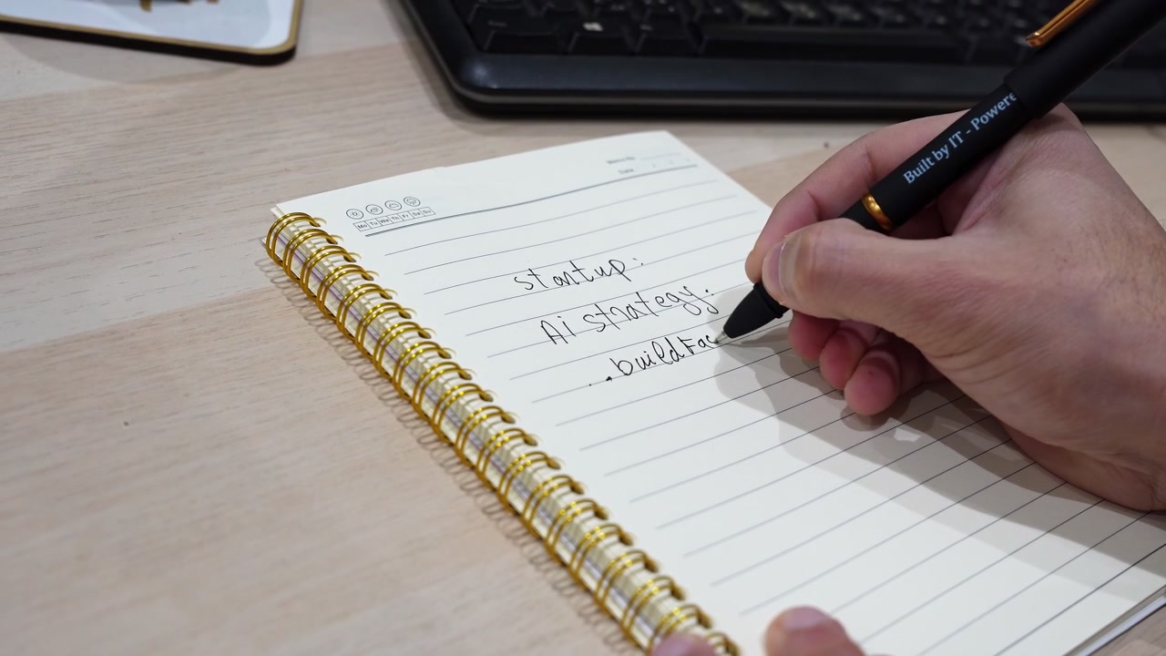 Close up shot of a male hand using a black pen to write an AI startup strategy in a spiral notebook. The handwritten notes about testing ideas sit on a wooden office desk near a computer keyboard.