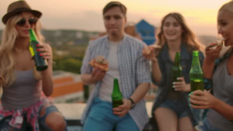 Las-Niñas-Y-Los-Niños-Rusos-Tintinean-Vasos-Y-Beben-Cerveza-De-Botellas-Verdes-En-La-Fiesta-Con-Amigos-En-El-Techo-Al-Atardecer.-Están-Sentados-Juntos-Comiendo-Pizza-Caliente-Después-De-Las-Noches-De-Verano.