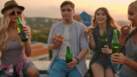 las chicas y los chicos rusos tocan vasos y beben cerveza de botellas verdes en la fiesta con amigos en el techo al atardecer. están sentados juntos comiendo pizza caliente después en verano.