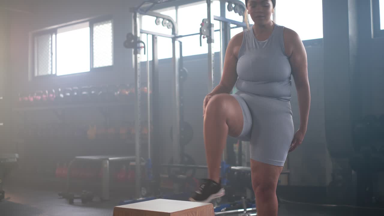 Donna afroamericana che si allaccia le scarpe da ginnastica blu, sale su una plyo box da palestra, afferra una bottiglia e si prepara per l'allenamento