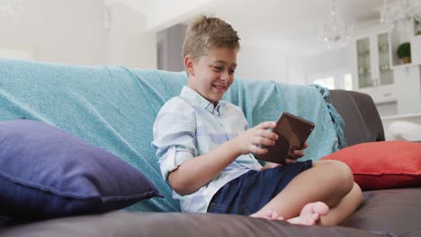 happy caucasian boy at home, sitting on couch using tablet, playing game and smiling