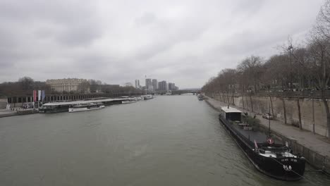 Toma-Estática-Del-Paisaje-Urbano-Del-Río-Sena-Que-Fluye-A-Través-De-La-Ciudad-De-París,-Barcos-Anclados-A-Un-Lado,-Distrito-Financiero-En-La-Distancia