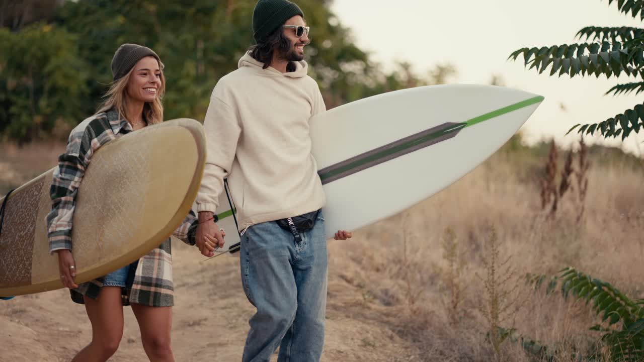 un chico feliz con gafas de sol en una sudadera blanca camina con su novia rubia en una camisa a cuadros con tablas de surf en sus manos a lo largo de un camino a lo largo del césped seco de verano y pequeños árboles