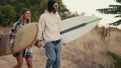 Un-Chico-Feliz-Con-Gafas-De-Sol-Y-Una-Sudadera-Blanca-Camina-Con-Su-Novia-Rubia-Con-Una-Camisa-A-Cuadros-Y-Tablas-De-Surf-En-Las-Manos-Por-Un-Sendero-Entre-La-Hierba-Seca-De-Verano-Y-Pequeños-árboles.