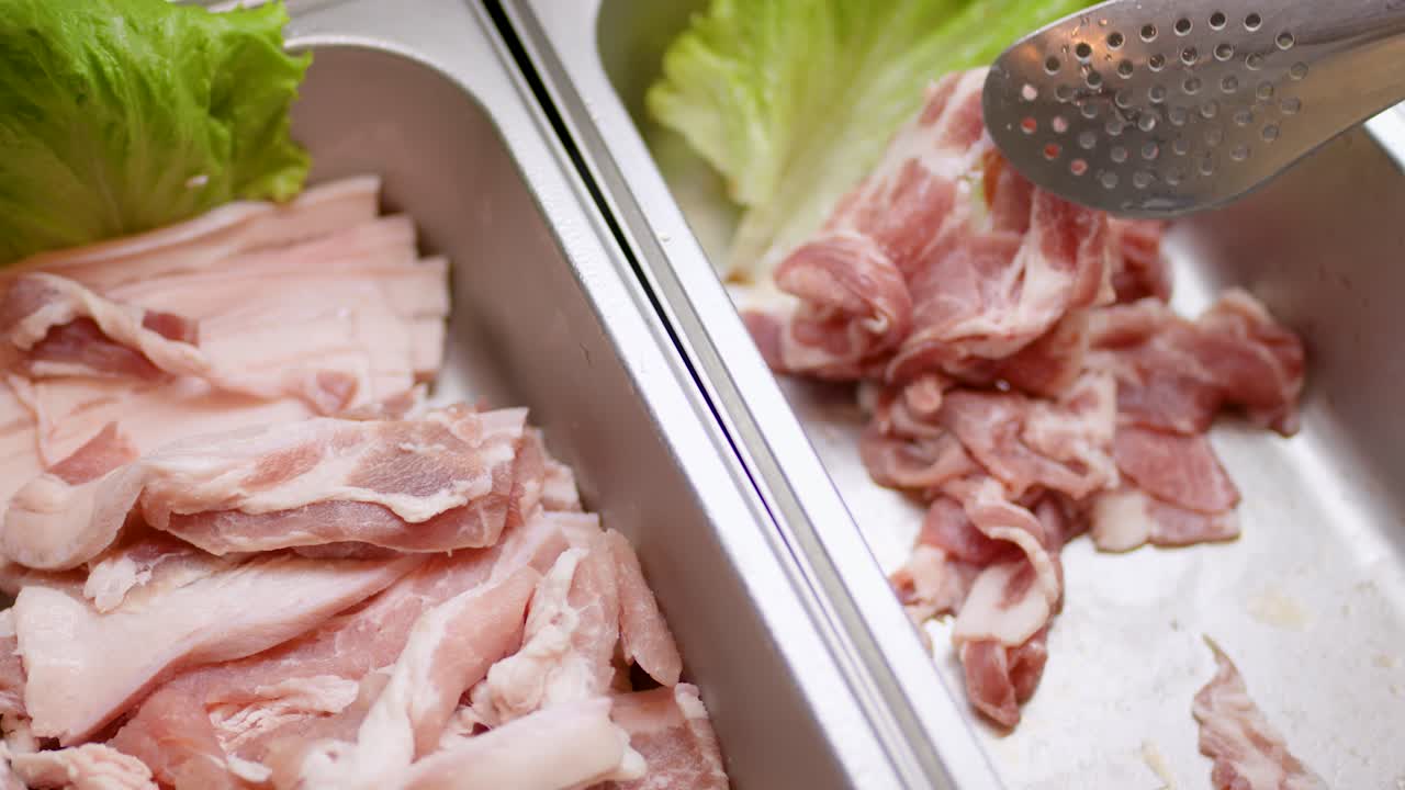 Hand uses metal tongs to pick raw pork slices from stainless buffet tray under lights