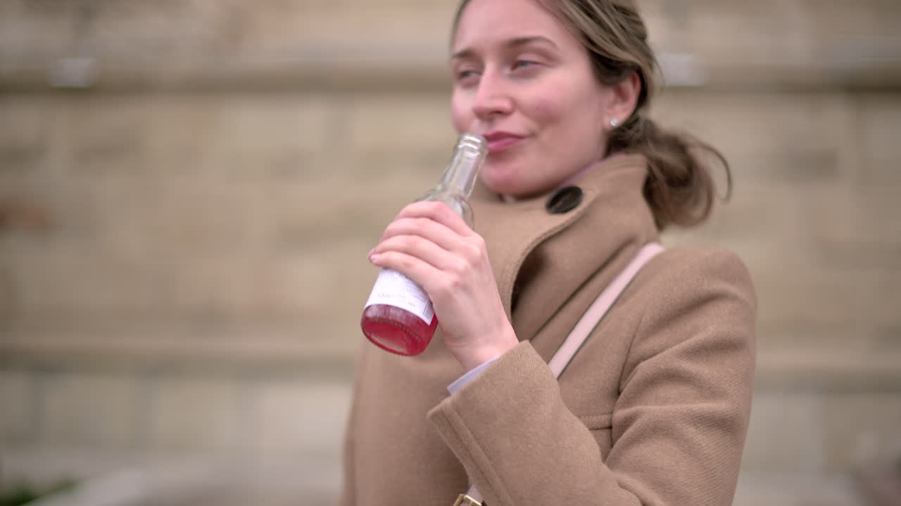 Woman in brown coat drinking a pink beverage outside