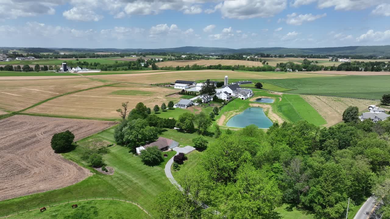 Aerial view of a picturesque American farm with lush green fields, serene pond and charming farm buildings. Rolling hills stretch into the distance under a bright blue sky. Peaceful rural landscape.