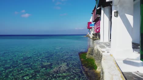 vista deslizante que muestra aguas poco profundas y guijarros bajo el agua a lo largo de las casas en el paseo marítimo de little venice en el casco antiguo de mykonos, grecia