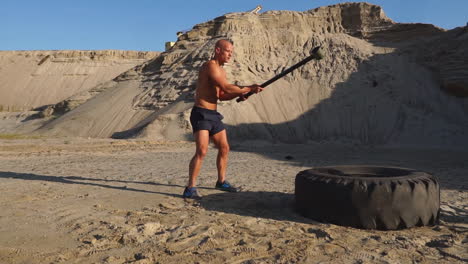 el hombre calvo golpea un martillo en una rueda enorme en las montañas arenosas en cámara lenta. entrenamiento de fuerza y resistencia para luchadores.